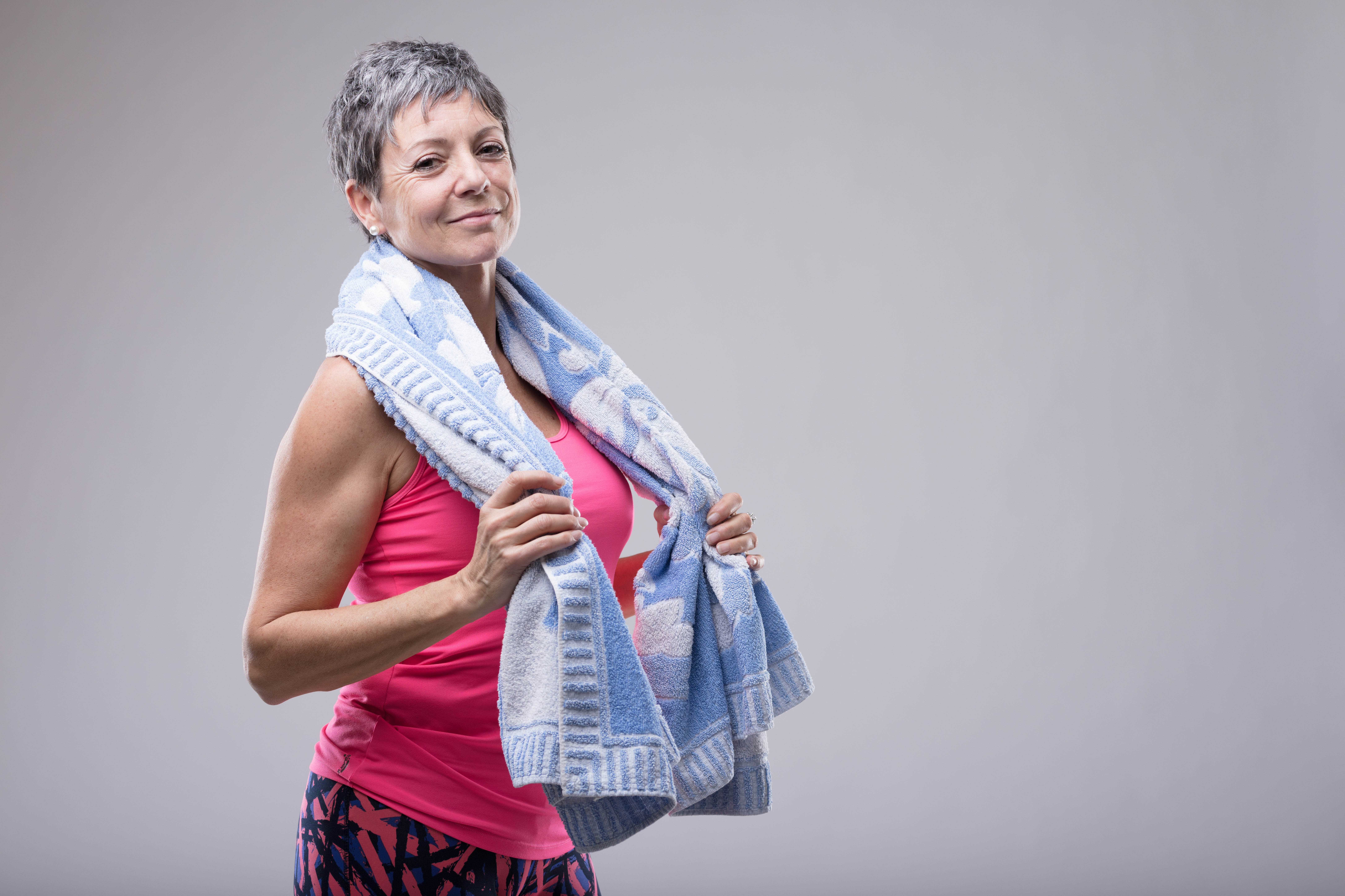 Older woman dressed in pink workout gear, standing with a towel wrapped around her neck, hands holding the towel, preparing for a workout Older woman dressed in pink workout gear, standing with a towel wrapped around her neck, hands holding the towel, preparing for a workout